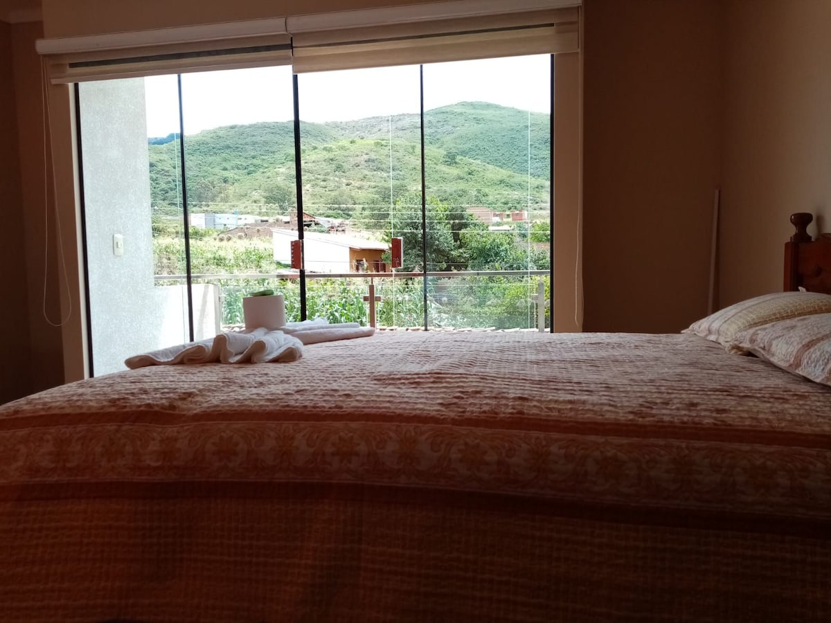 A spacious bedroom features a large bed with a patterned blanket. Floor-to-ceiling windows offer views of green hills and distant structures. Natural light filters in, enhancing the serene atmosphere of the room.