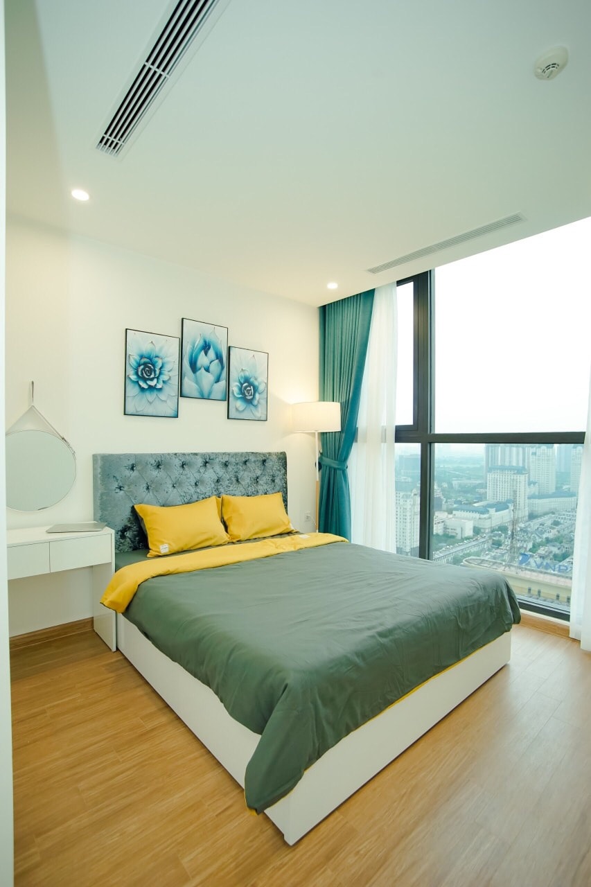 A cozy bedroom features large windows offering city views. A plush bed is neatly dressed with a grey comforter and bright yellow accents. Blue wall art complements the decor, while a light fixture and a small vanity enhance the space's functionality.