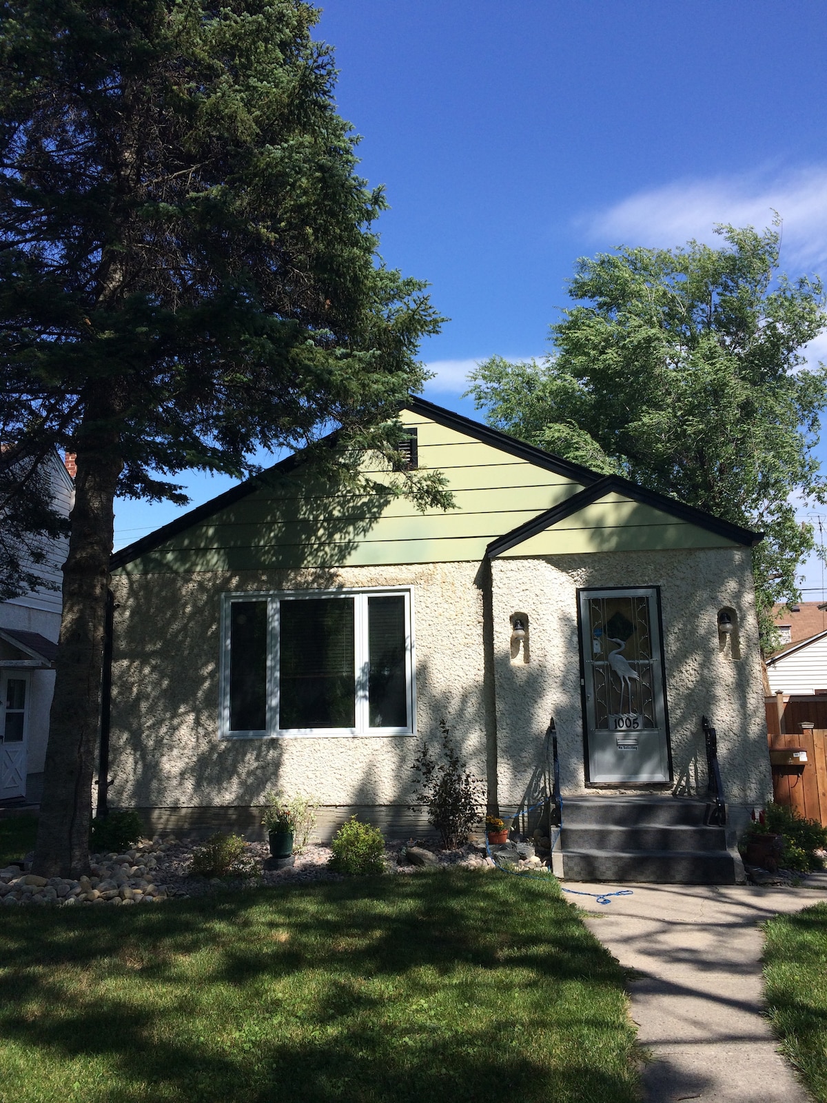The exterior of the bungalow features a light green gabled roof and a stone facade. Large front windows provide ample natural light. A well-maintained lawn with shrubs and trees enhances the inviting entrance, complemented by a stone pathway leading to the front door.