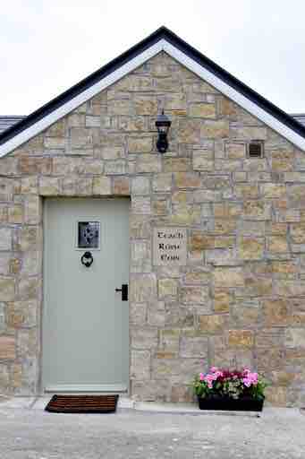 The entrance of a stone cottage features a pale green door adorned with a decorative window. A small potted plant with colorful flowers is positioned beside the door, enhancing the welcoming appearance of the home.