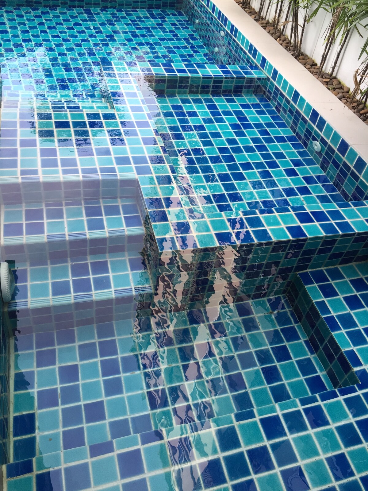 A vibrant swimming pool area features a mosaic of blue and green tiles, reflecting light across the surface of the water. Steps lead down to a sunken section, creating an inviting space for relaxation.