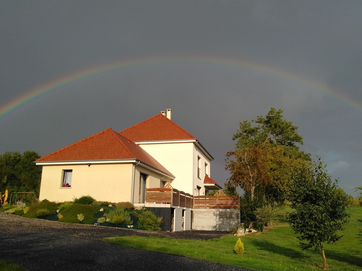 A charming house features a sloped roof and a covered deck, surrounded by well-maintained greenery. A rainbow arches gracefully in the sky above, contrasting with the clouds. The property is set in a tranquil landscape, offering a peaceful and picturesque view.