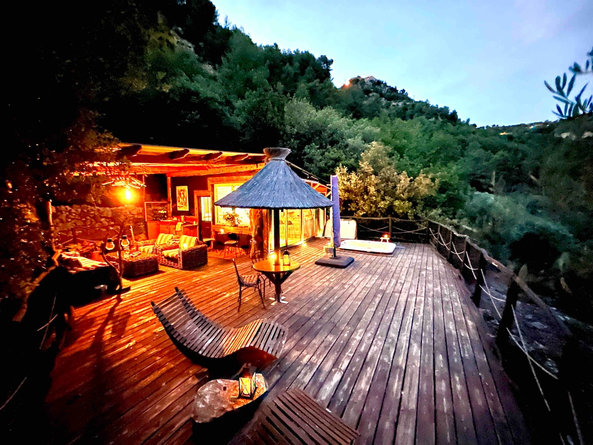 A wooden terrace features comfortable seating arrangements, a large parasol, and a hammock, surrounded by lush greenery. Soft lighting illuminates the outdoor space, creating an inviting ambiance as evening falls. A serene atmosphere is suggested by the natural surroundings.