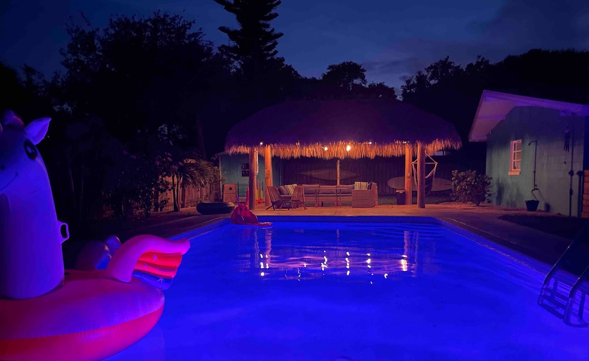 The pool area is illuminated with soft blue lighting, casting a calming glow on the water. A tiki hut is visible in the background, featuring comfortable seating and tropical decor. Colorful pool floaties add a fun touch to the inviting atmosphere.