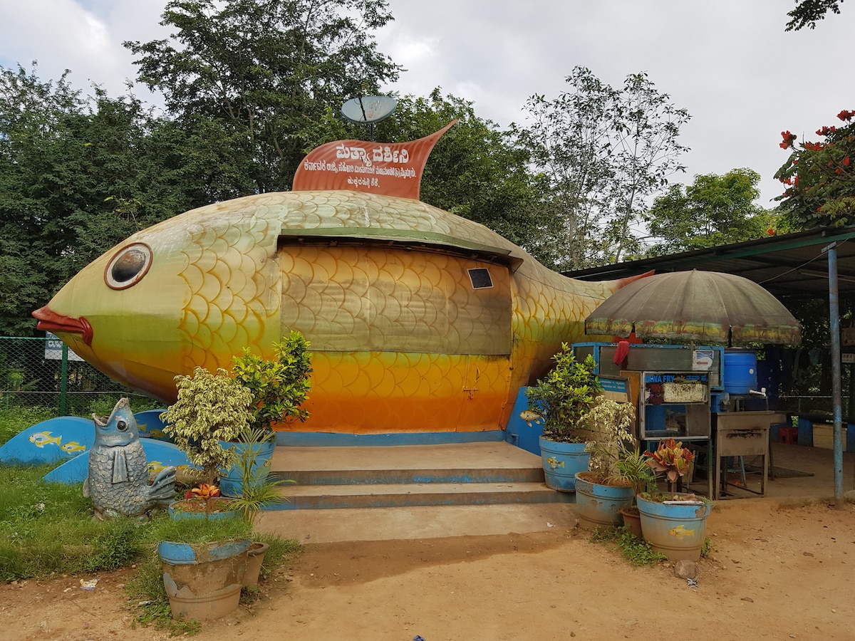 A whimsical structure resembling a large fish is set in a natural environment. The fish-shaped building features an open doorway and a vibrant, multicolored exterior. Surrounding plants and pots add greenery, while a small kiosk is positioned nearby, offering refreshments and seating under an umbrella.
