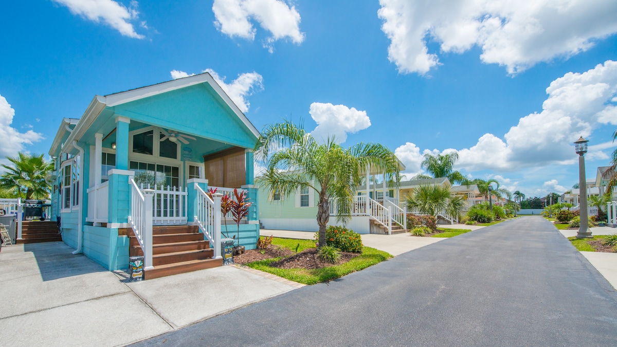 A charming manufactured home is showcased with a bright blue exterior, flanked by palm trees and lush landscaping. A welcoming porch is featured, leading to spacious steps. A clean, paved pathway continues past the home, lined with similar residences under a clear blue sky.