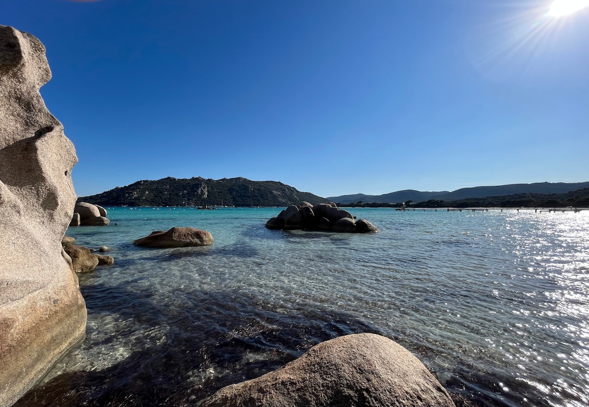 A serene coastal view features smooth, large rocks at the water's edge, surrounded by clear, turquoise waters. The landscape includes distant hills under a clear blue sky, with sunlight reflecting off the water's surface, creating a tranquil atmosphere.
