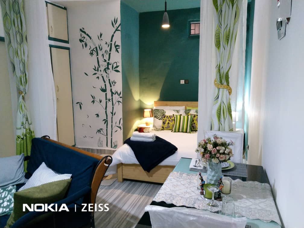 A cozy room is adorned with green and white accents, featuring a spacious bed with various patterned pillows. A small dining table with a vase of flowers is positioned in the foreground. Light streams through sheer curtains, illuminating the bamboo-patterned wall design.