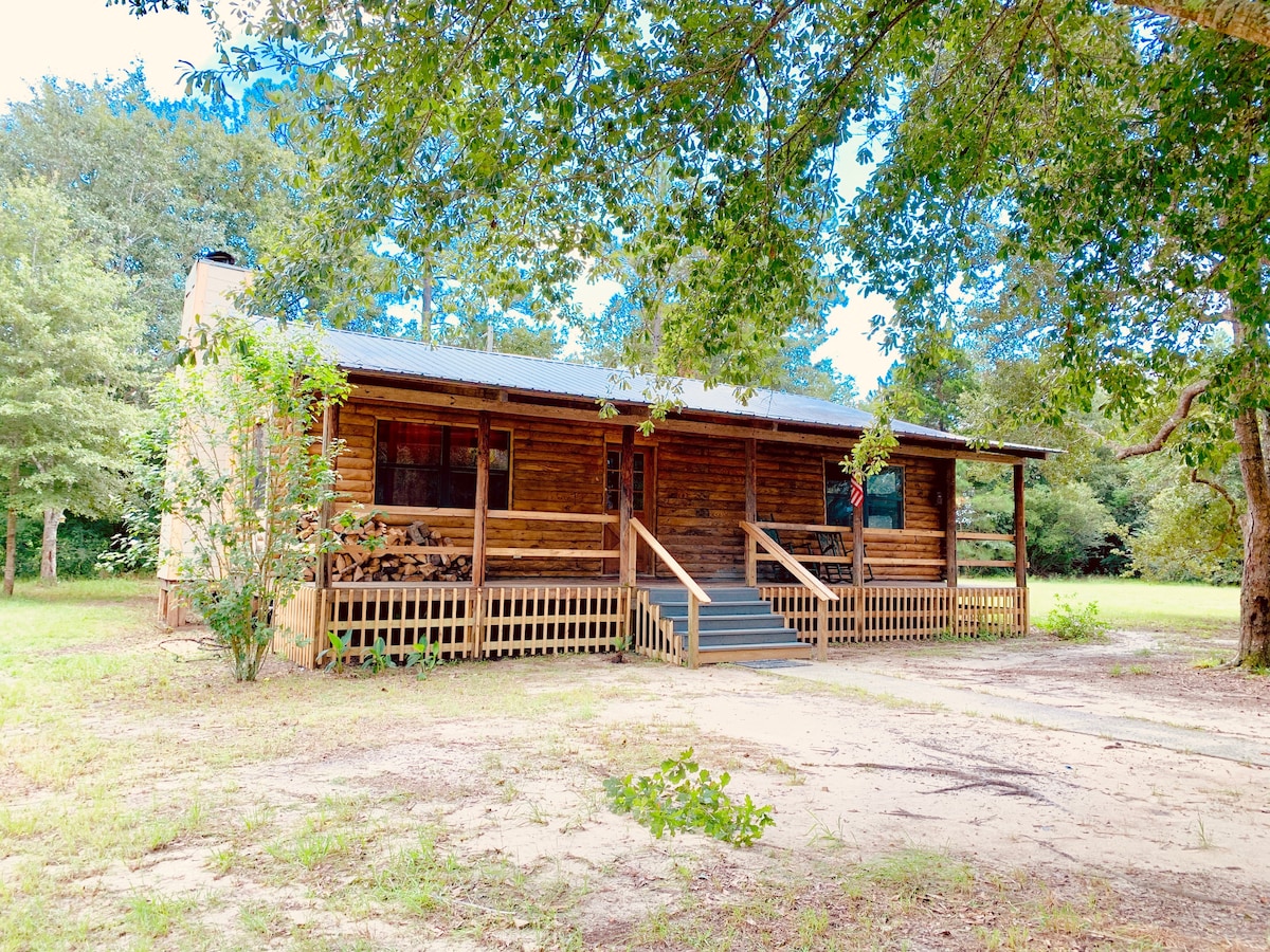 A rustic log cabin is surrounded by lush greenery, featuring a front porch with steps leading up to the entrance. The structure blends with the natural landscape, showcasing wooden elements and a metal roof.