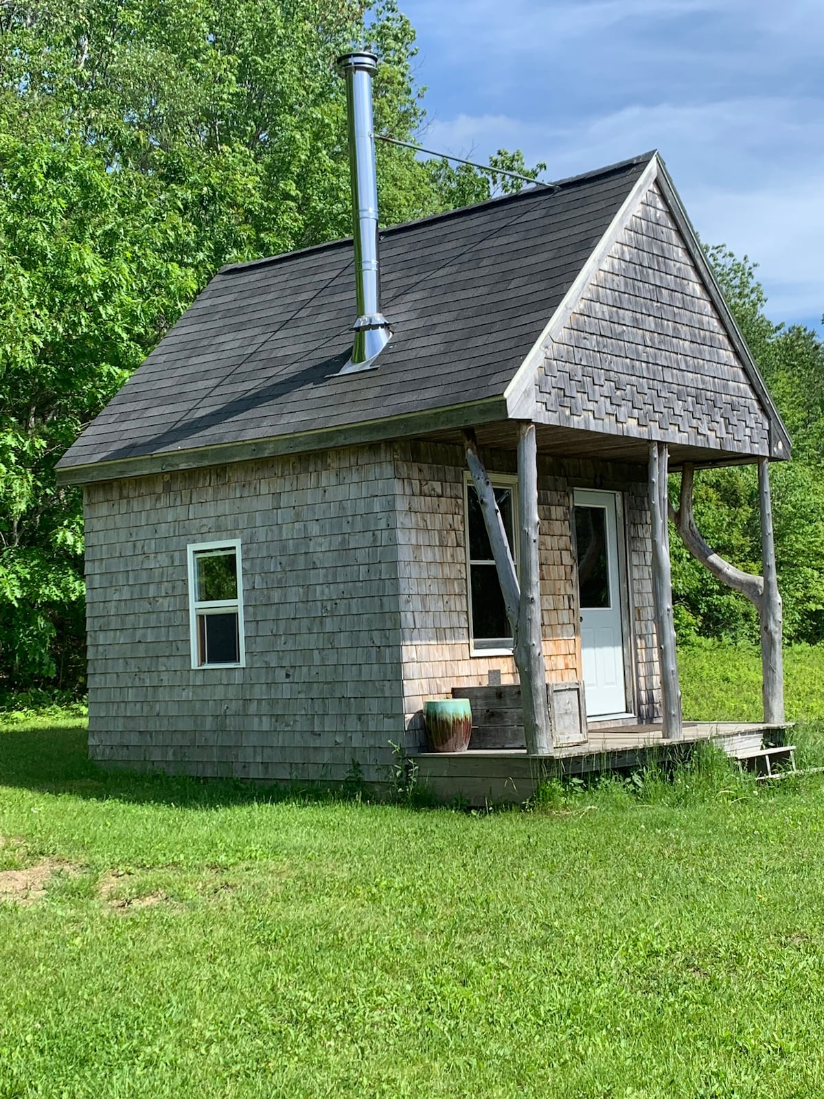 A small, rustic cabin is positioned in a grassy area, surrounded by trees. The cabin features wooden shingles and a pitched roof, with a chimney extending from the top. A white door is visible, flanked by a single window. A small porch area leads to the entrance.