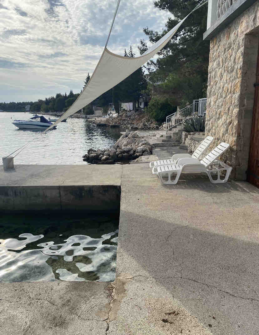 A stone-finished area extends towards a serene waterfront, featuring two white lounge chairs positioned on the edge. A small boat is seen moored in the calm water, while trees line the shore in the background. A sunshade is stretched overhead.