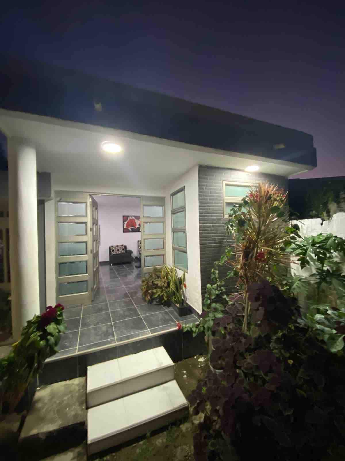 The entrance of a modern accommodation is showcased, featuring a small set of white stairs leading up to glass-paneled doors. Lush greenery surrounds the pathway, and subtle outdoor lighting illuminates the space in the evening twilight.