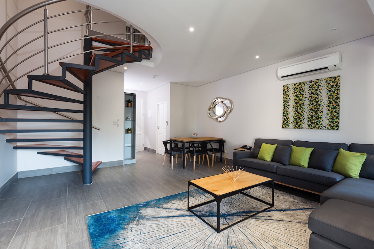 A modern living area features a spiral staircase leading to upper levels. A navy sectional sofa with green accent pillows is positioned around a coffee table. A unique wall art piece and a dining table create a balanced space under soft lighting.