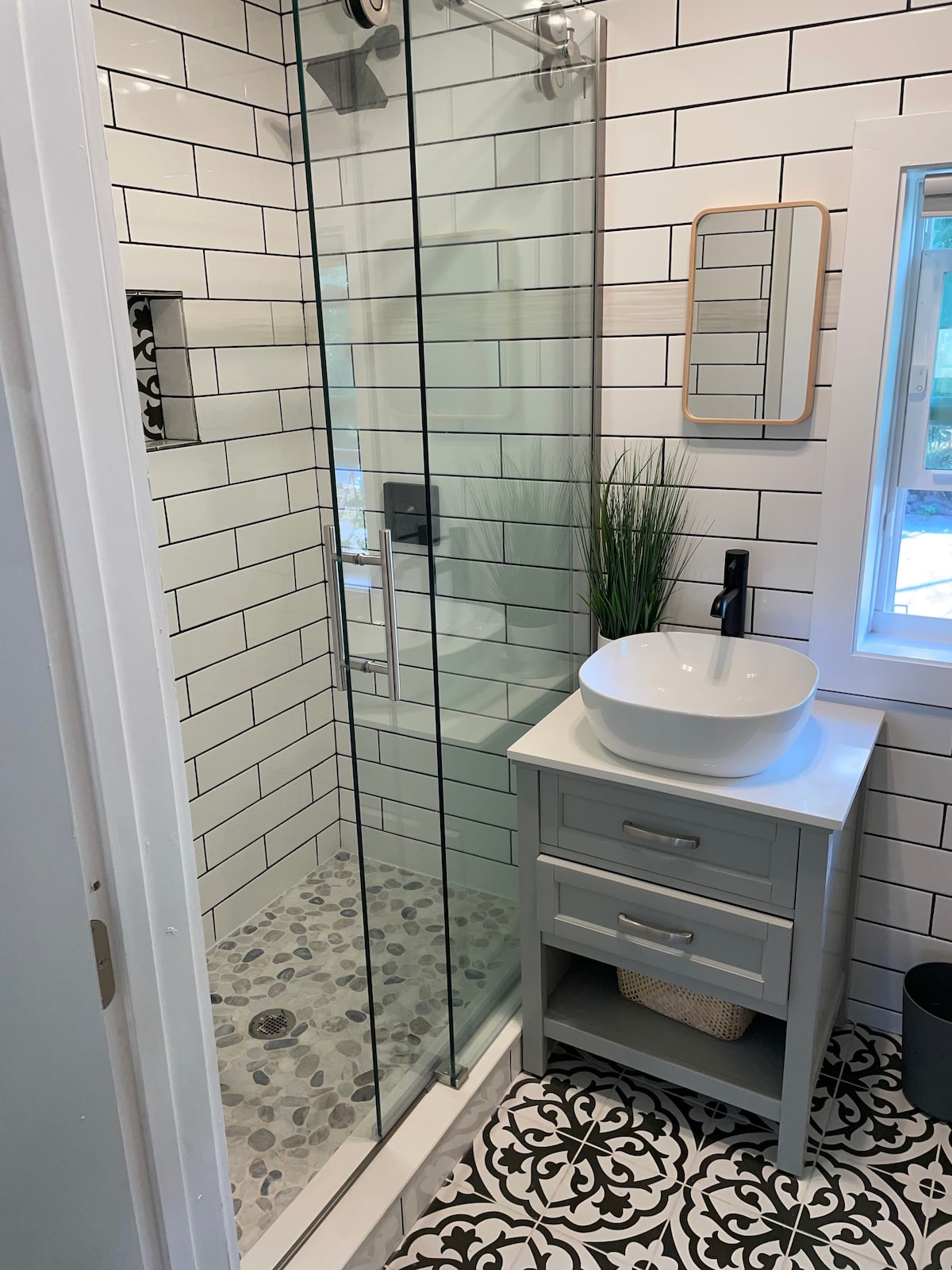A contemporary bathroom features a glass shower with a pebble stone floor and sleek black fixtures. A minimalist sink is set atop a gray cabinet with two drawers. A round mirror hangs above the sink, and a window provides natural light, accented by decorative black and white tile flooring.
