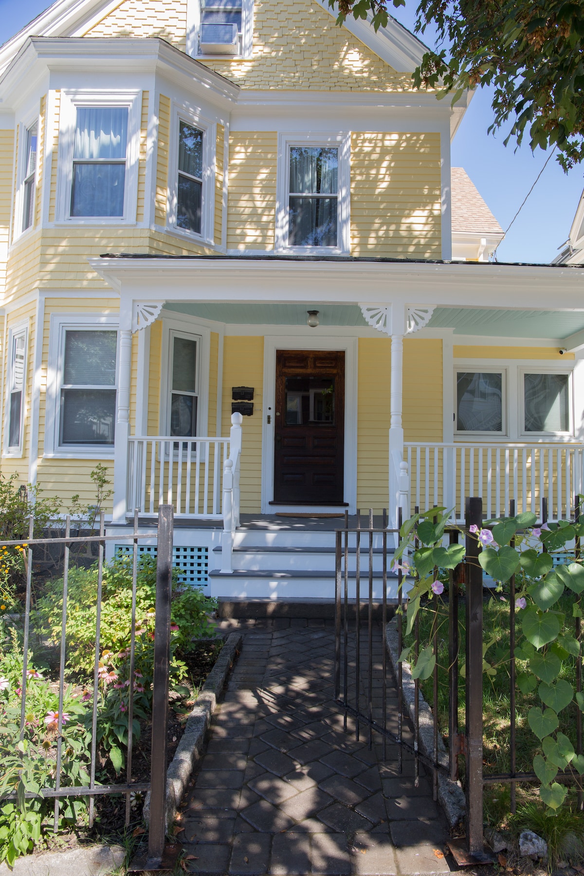 The exterior of the yellow house features a welcoming front porch adorned with white railings and decorative accents. A pathway made of stone leads from the wrought iron gate to the front door, surrounded by well-maintained garden beds with greenery and colorful flowers.