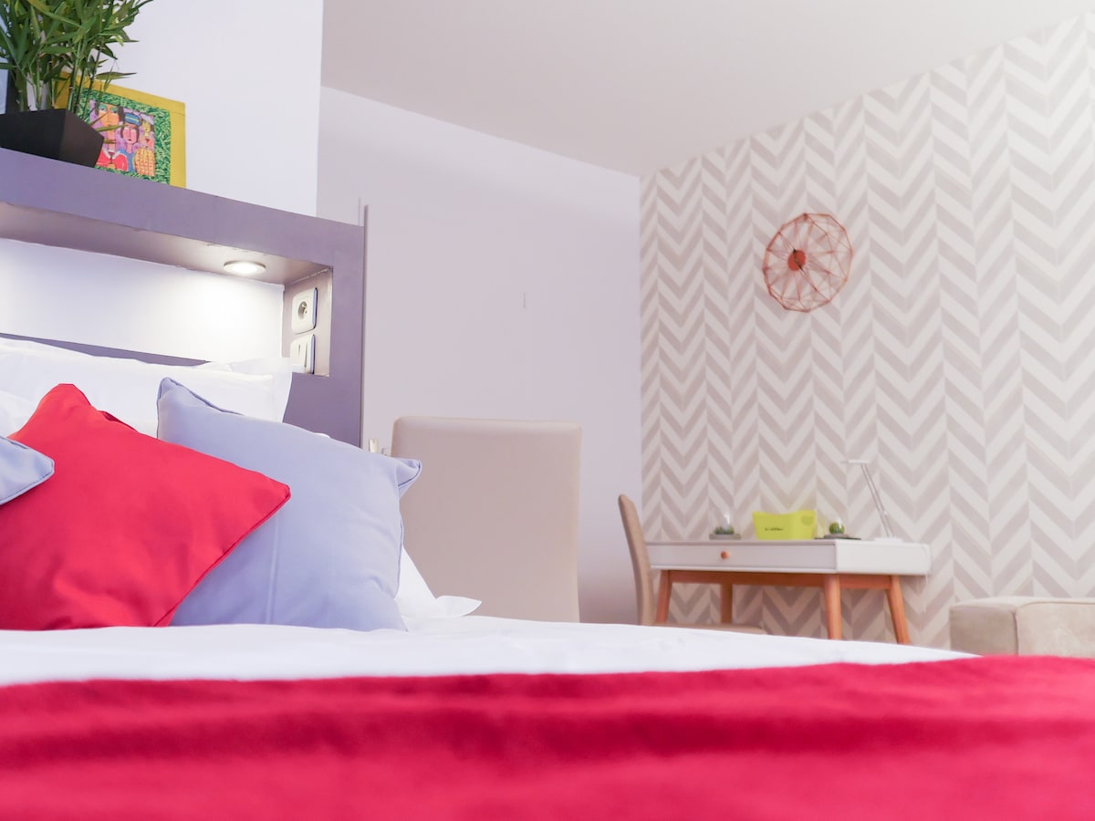 A spacious bed is adorned with soft pillows in shades of red and blue. The background features a light-colored wall with a geometric pattern and a small desk positioned nearby. Natural light from an unseen window brightens the inviting space.