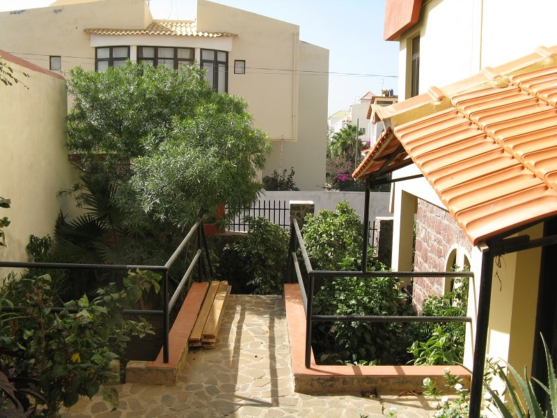 A pathway is framed by lush greenery and plants, leading towards a secluded area within the property. Wooden benches are placed along the walkway, and a structure with a terracotta roof is seen in the background, contributing to a peaceful outdoor setting.