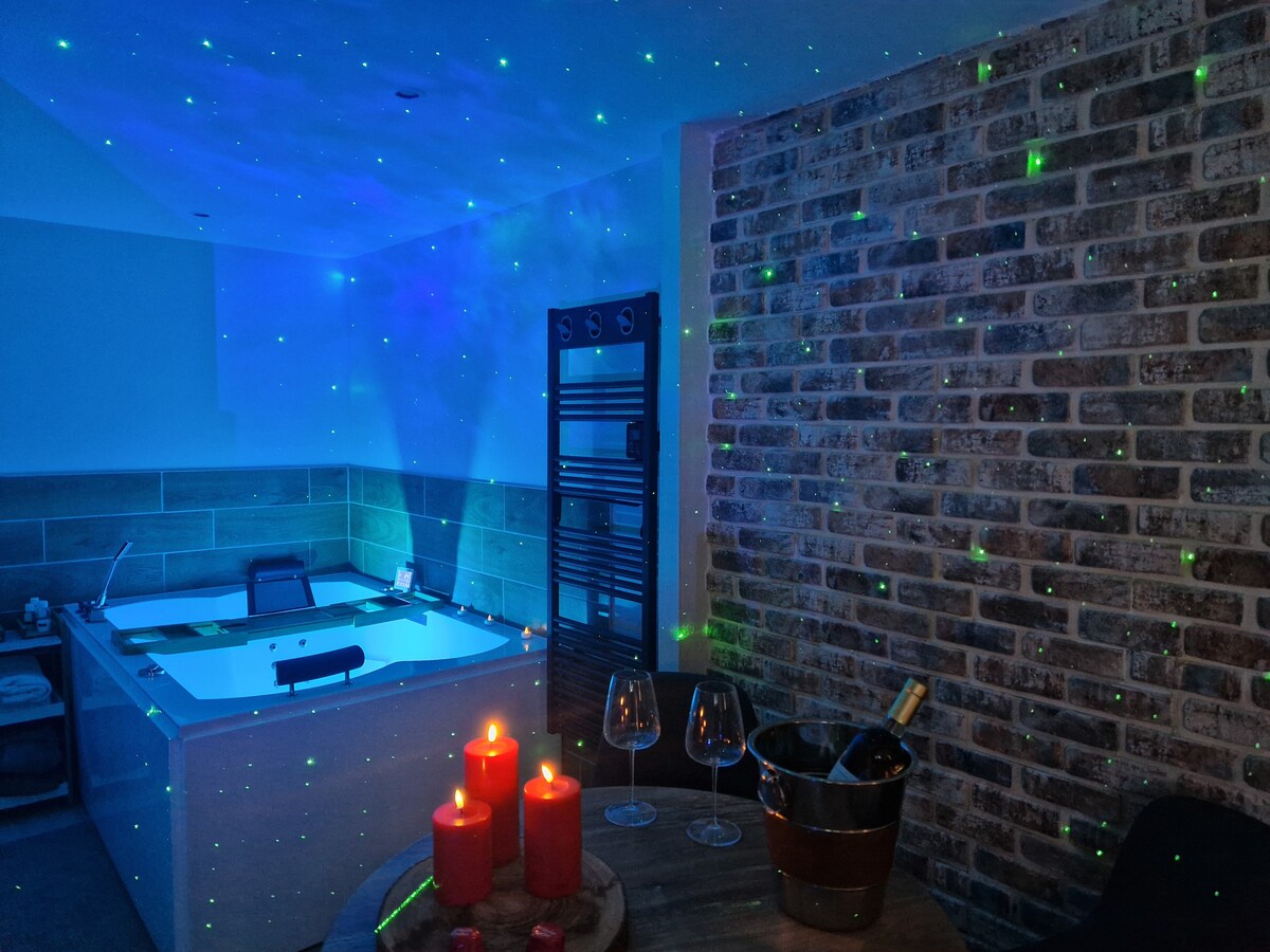 A spa area is illuminated by soft blue lights, creating a calming atmosphere. A large soaking tub is visible alongside a stylish heated towel rack and a textured brick wall. A small table features a wine bucket and elegantly placed glasses, with lit candles adding warmth.