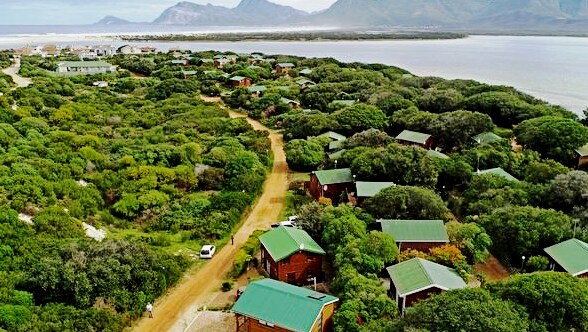An aerial view showcases a serene landscape dotted with cozy cabins surrounded by lush greenery. A winding dirt road leads through the area, with views of mountains and the lagoon in the background, highlighting the tranquil setting of the Middelvlei Reserve.