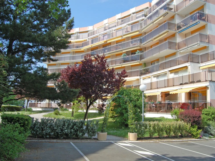 The image depicts a multi-story residential building with a curved façade. Lush greenery, including trees and flowering shrubs, surrounds the entrance. Balconies are visible on each floor, enhancing the building's modern design. Parking spaces are located in the foreground, providing accessible options.