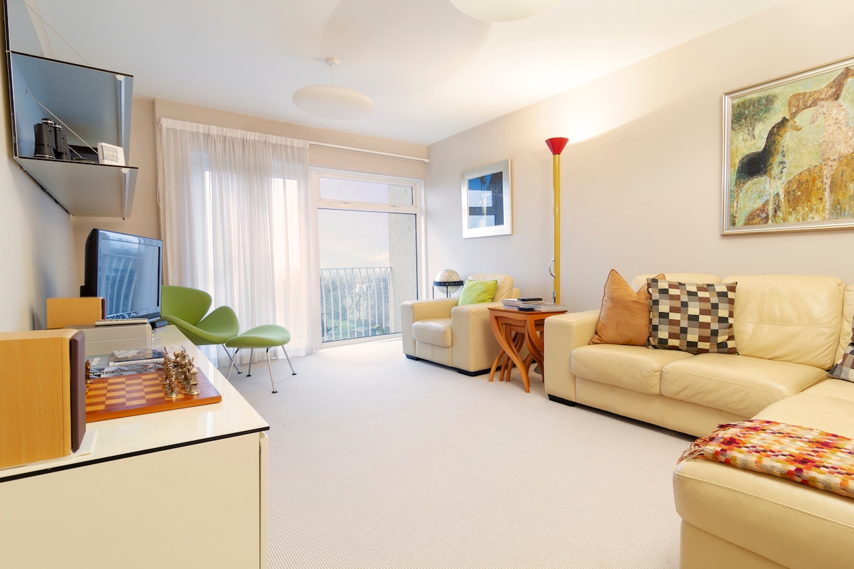 The living area is furnished with light-colored sofas, a patterned throw, and a sleek coffee table. A television and a decorative shelf are positioned against the wall. A floor lamp adds illumination, and a sliding glass door leads to a small balcony, revealing views of the surroundings.
