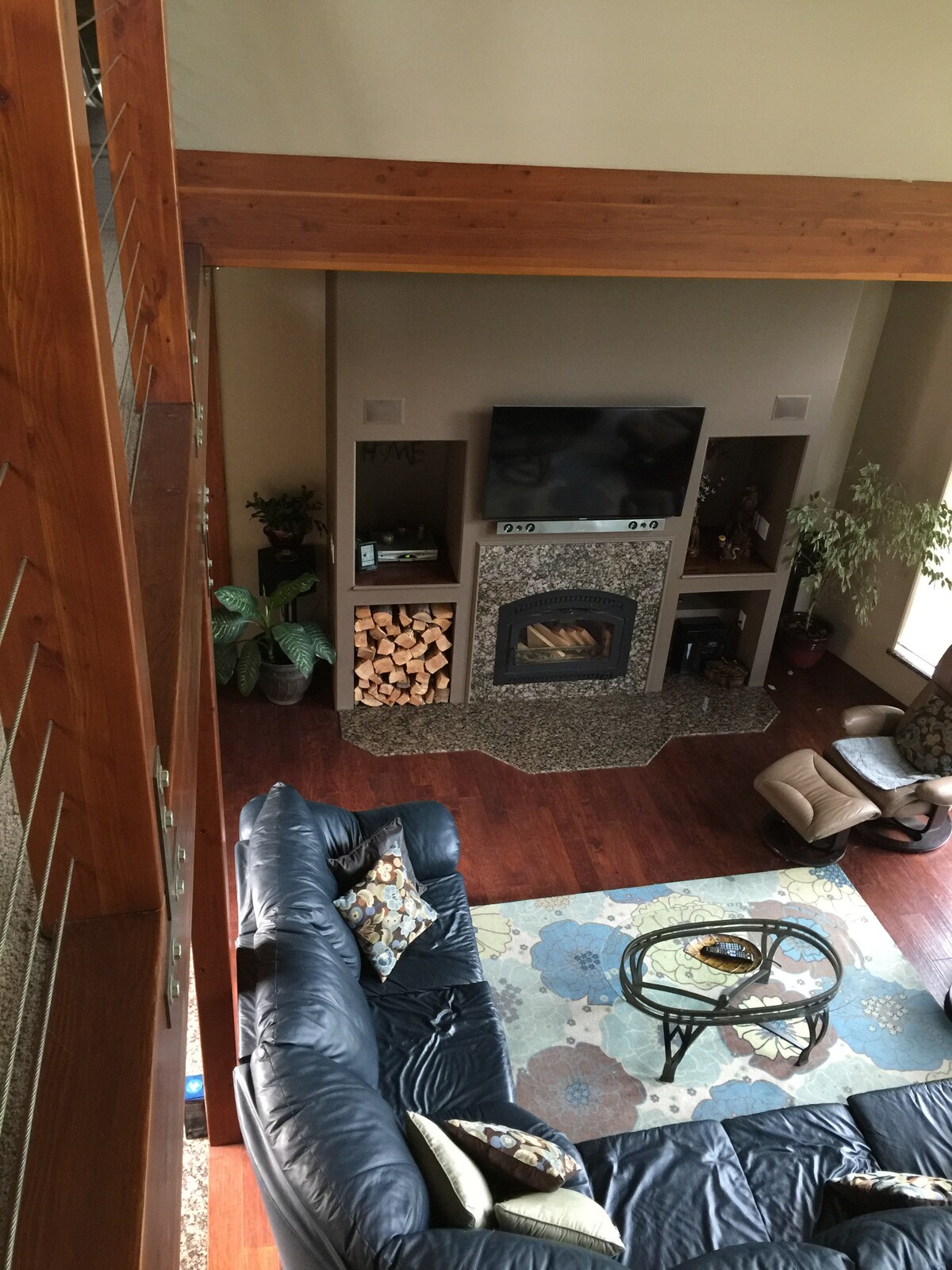 An open living space features a leather sectional sofa, a patterned area rug, and a glass coffee table. A media console holds a flat-screen television, while wood logs are stacked beside a stone fireplace. Decorative plants add greenery to the room.