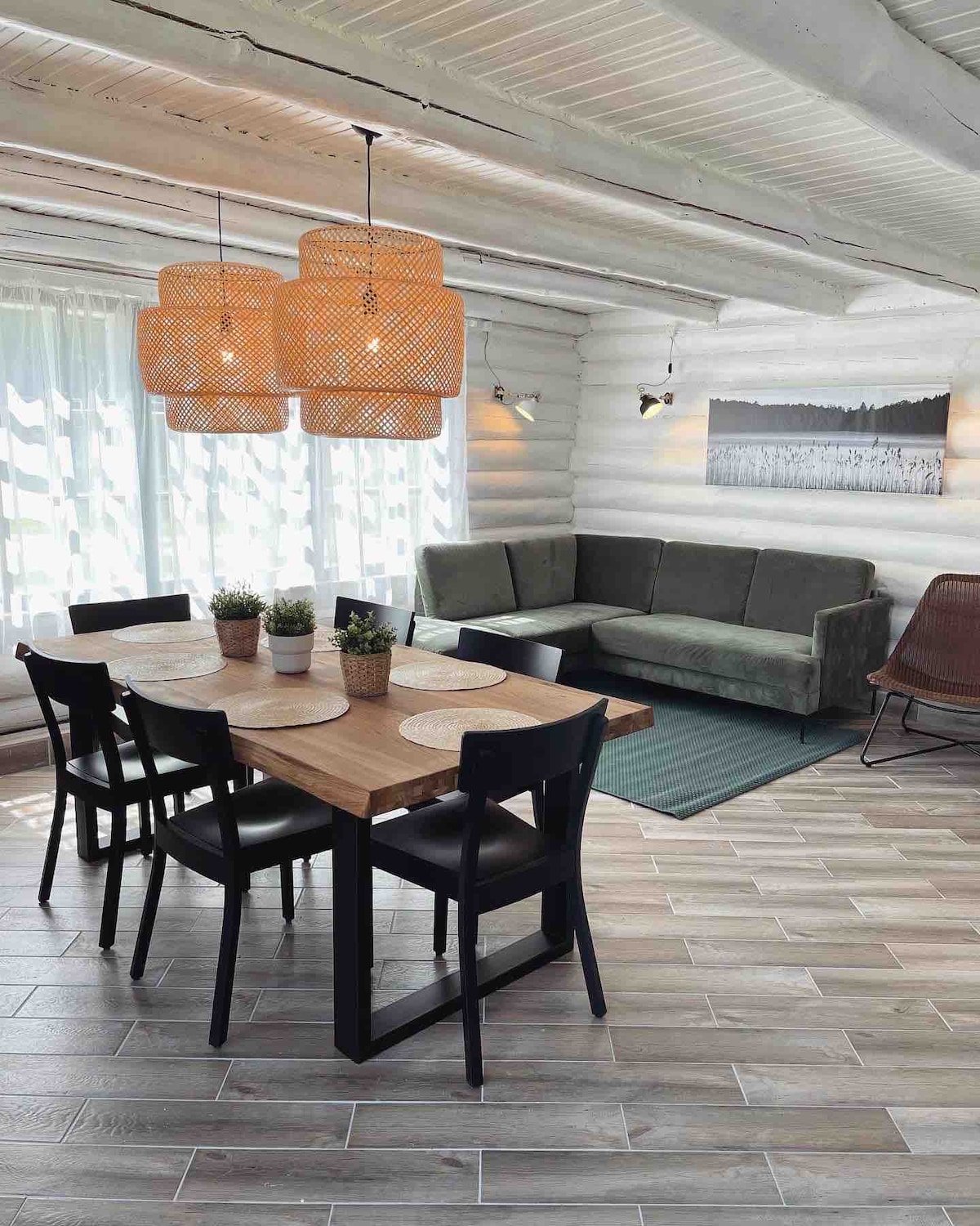 A dining area features a wooden table surrounded by six black chairs. A cozy green sofa is positioned nearby, complementing light-colored walls and soft natural light streaming through sheer curtains. Two woven light fixtures add texture overhead, enhancing the modern design.