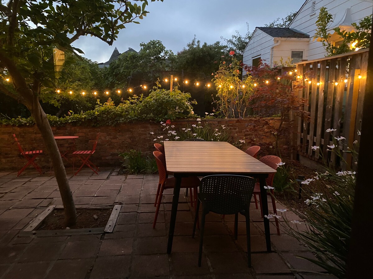 An outdoor patio is illuminated by warm string lights, surrounded by vibrant greenery and flowering plants. A large table with colorful chairs is center stage, complemented by smaller tables set against the brick wall. The serene atmosphere invites relaxation and outdoor dining.