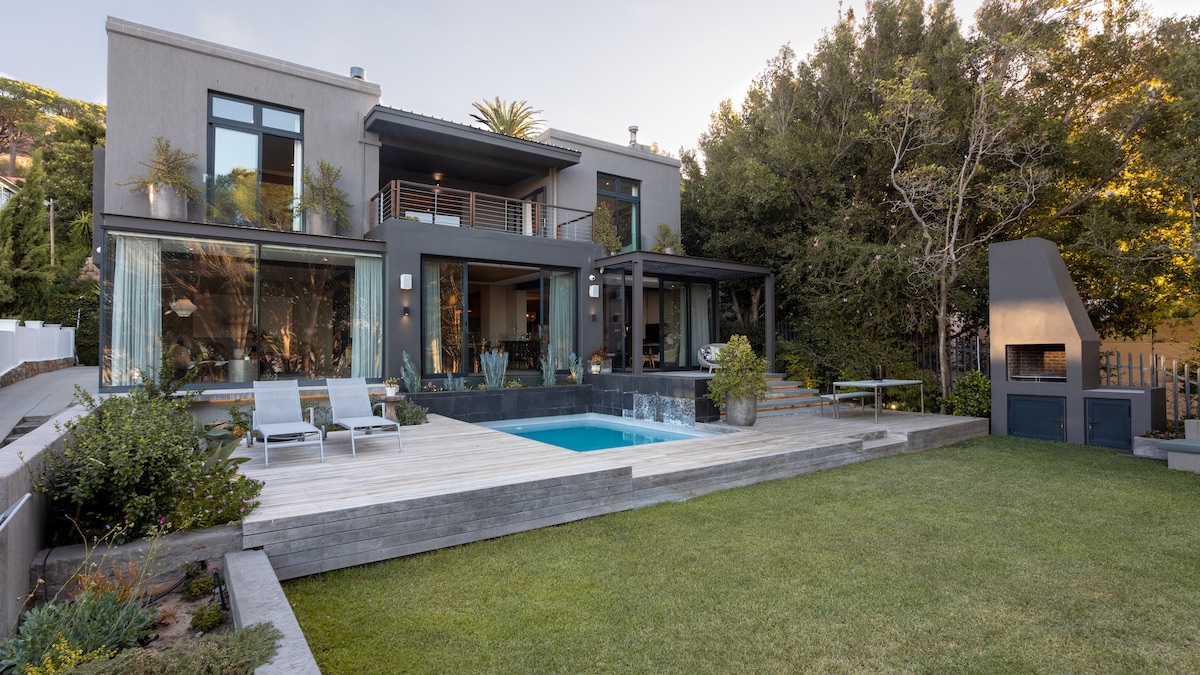 A modern villa is surrounded by lush greenery, featuring a swimming pool and a wooden deck with loungers. Large glass doors provide an unobstructed view of the outdoor space, which includes a landscaped garden and a built-in outdoor fireplace.