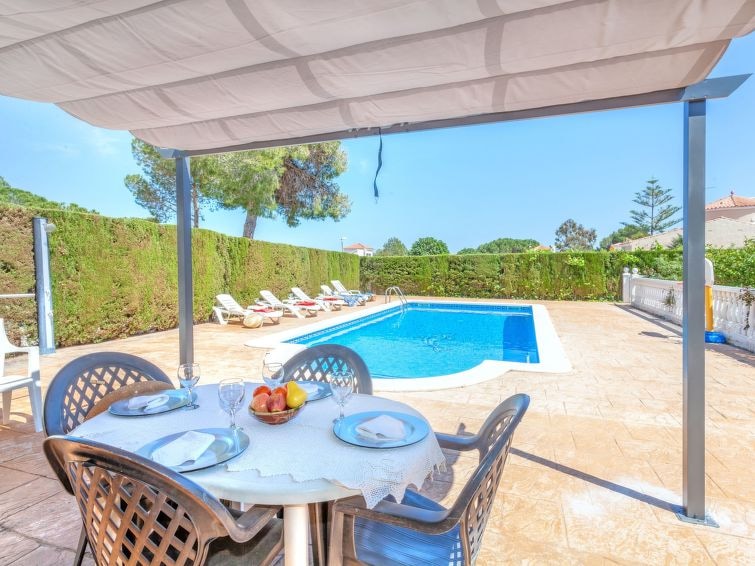 A covered dining area features a round table set for four, with fresh fruit displayed in the center. In the background, a rectangular swimming pool is framed by hedges and sun loungers, under a clear sky. The spacious outdoor space invites relaxation.