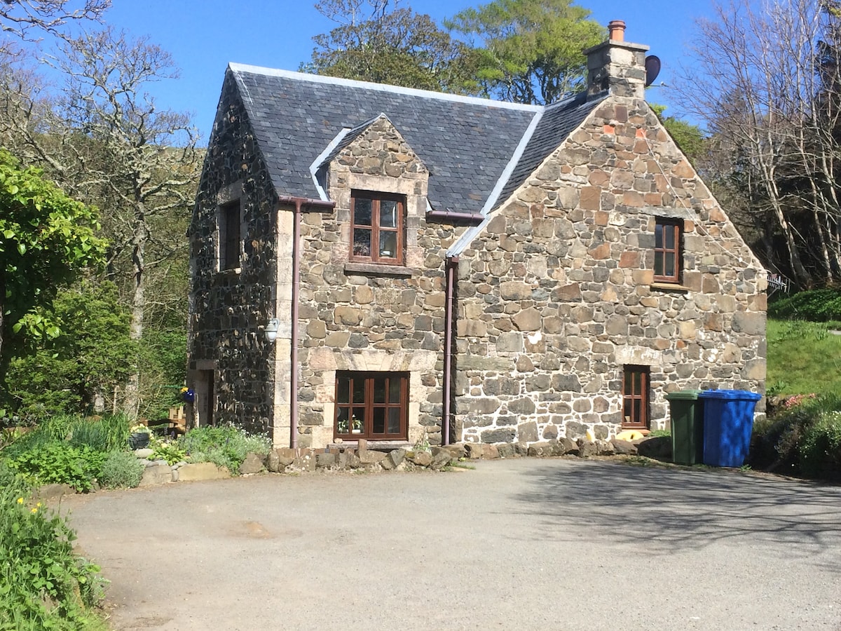 The rustic exterior of the stone house reflects traditional architecture, surrounded by lush greenery. The entrance features large windows, while the driveway provides access to the front of the home. A blue bin is positioned next to the building, adding to the site's charm.