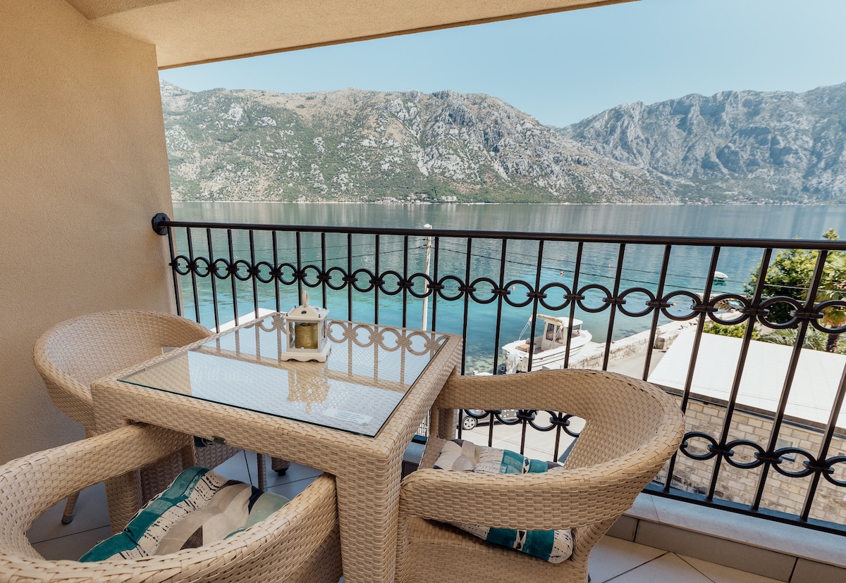 A balcony is depicted with a glass-top table surrounded by two woven chairs. The railing frames a panoramic view of the serene sea and mountains, emphasizing the natural beauty of the surrounding landscape.