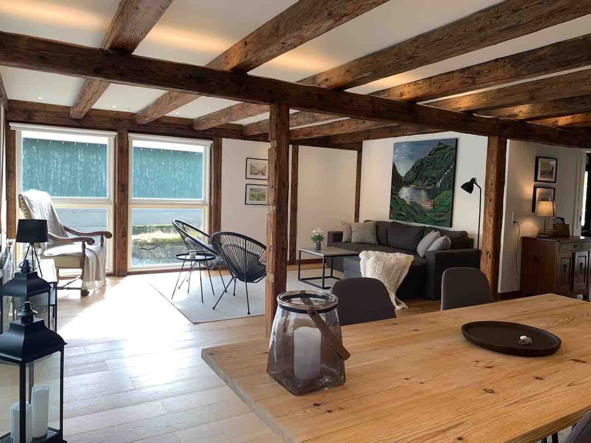The spacious living area features original exposed beams and wooden flooring. Large windows provide natural light and a view of the waterfront. The seating area includes a sofa and lounge chairs, complemented by a dining table, decorative art, and a lantern for ambiance.