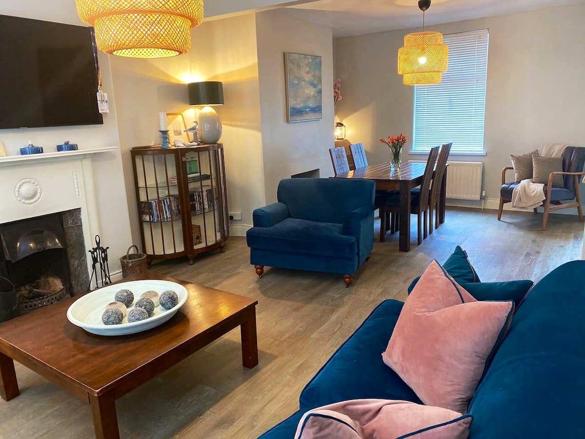 A spacious open-plan living area features a blue velvet sofa with plush pillows and a wooden coffee table displaying decorative spheres. A dining table sits to one side, accompanied by a cabinet containing books. Large windows allow natural light to fill the space, creating a warm environment.