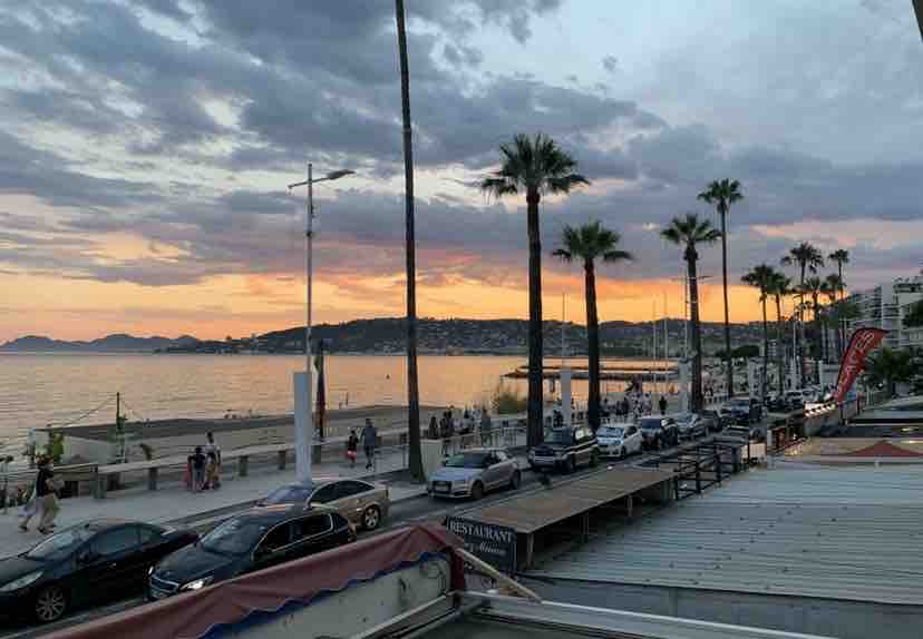 A vibrant beach scene is captured at sunset, with palm trees lining the promenade. The calm water reflects shades of orange and purple as people stroll along the shore. Vehicles are parked alongside the waterfront, creating an inviting atmosphere near the coastline.