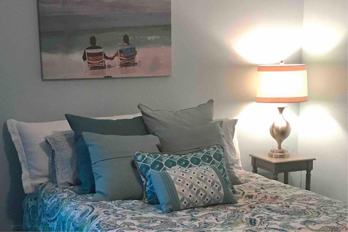 A comfortable sleeping area features a patterned bedspread adorned with multiple decorative pillows. A soft glow emanates from a bedside lamp, highlighting the adjacent artwork of two figures seated on a beach. The walls are painted in a calming light blue hue.