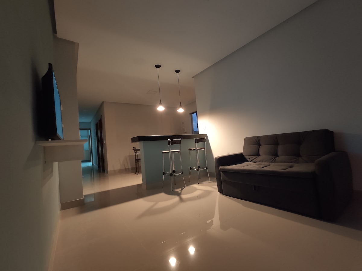 An open living space is depicted with a dark sofa positioned against a light wall. A modern kitchen counter with two bar stools is visible in the background. Soft pendant lighting illuminates the area, while glossy tiles reflect the light, creating a bright atmosphere.
