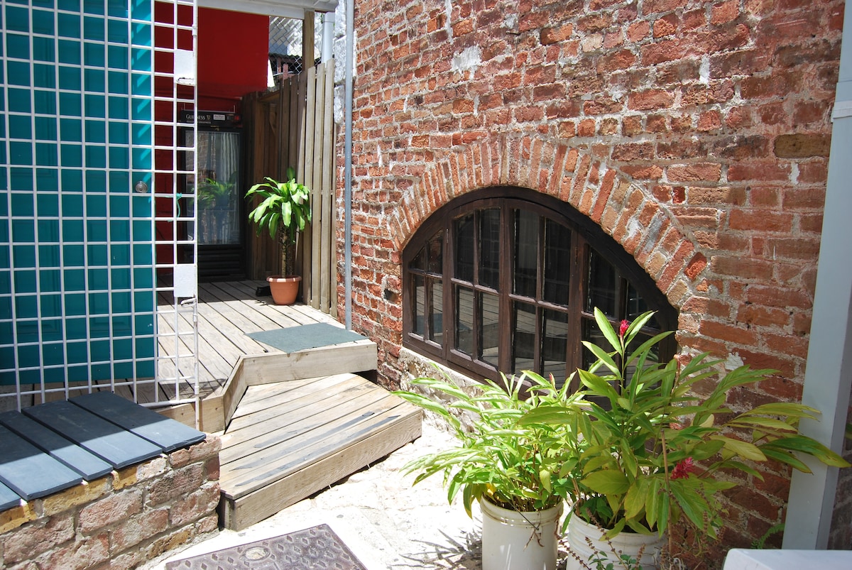 A charming courtyard area features a mix of brick and wooden elements. Lush greenery is positioned in a planter, adding vibrant color to the space. The brick wall showcases a rounded window, while a gentle slope leads from the wooden deck to the entrance.
