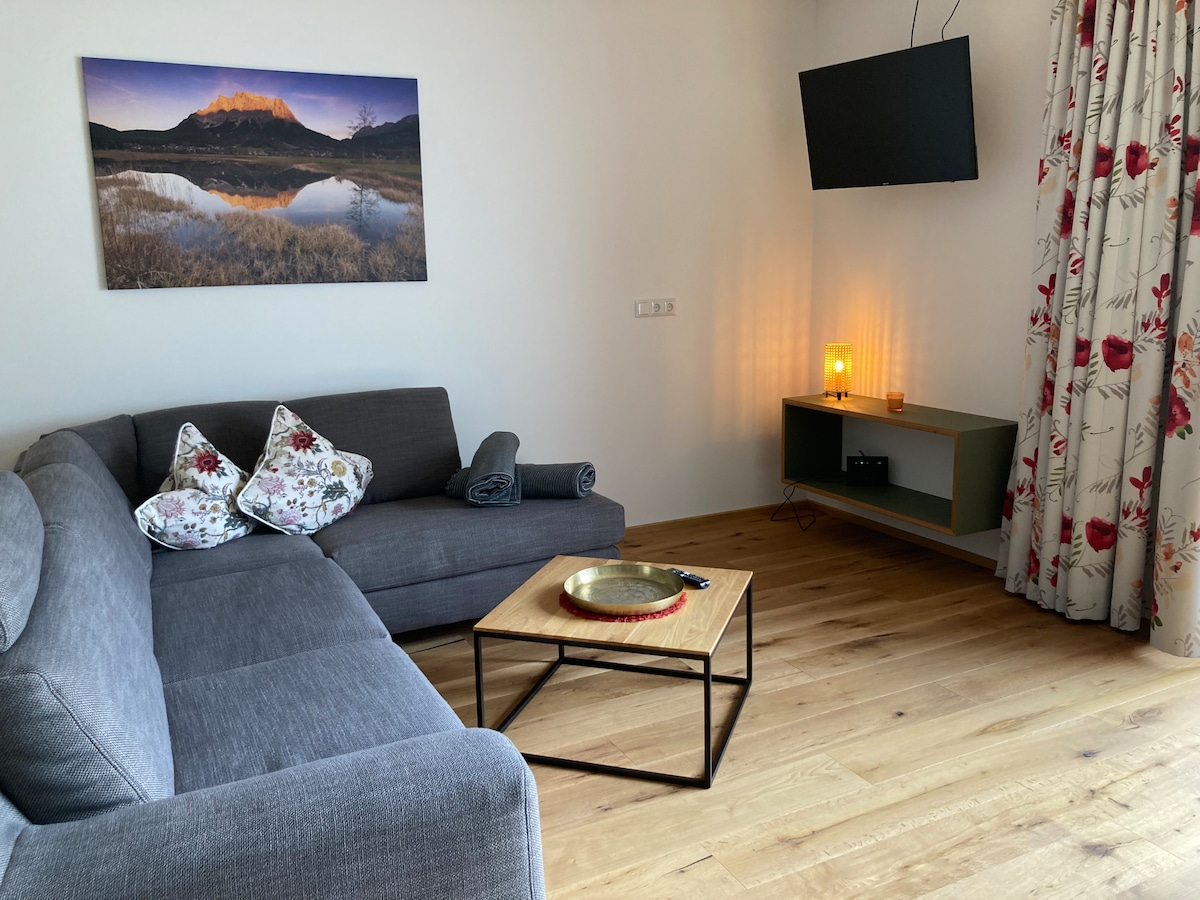 A comfortable living space is presented, featuring a gray sectional sofa with decorative pillows. A wooden coffee table sits at the center, with a TV mounted on the wall. Natural light illuminates the room, complemented by floral-patterned curtains and a soft glow from the lamp.