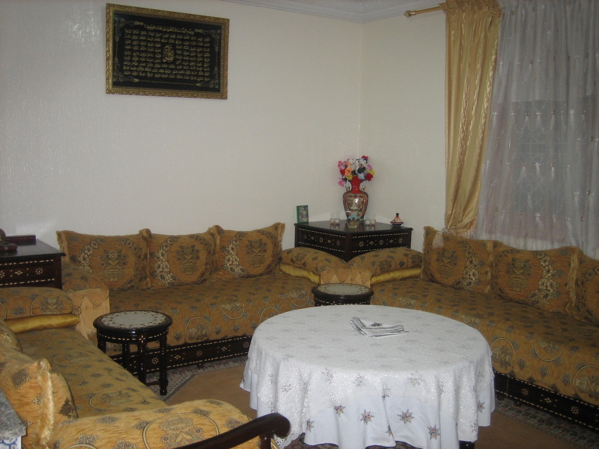 A cozy sitting area features intricately designed cushions arranged around a round table. Decorative side tables complement the seating, while a beautifully framed piece of artwork hangs above. Soft curtains filter light from a window, enhancing the room's warm ambiance.