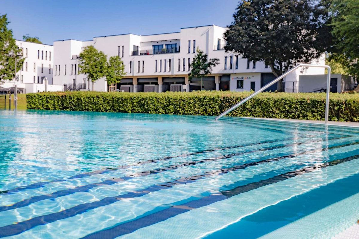 A serene outdoor pool is framed by neatly trimmed hedges and modern architecture. The water is clear and inviting, featuring subtle lane markings on the pool's surface. Soft sunlight reflects off the water, enhancing the calm atmosphere of the surrounding area.