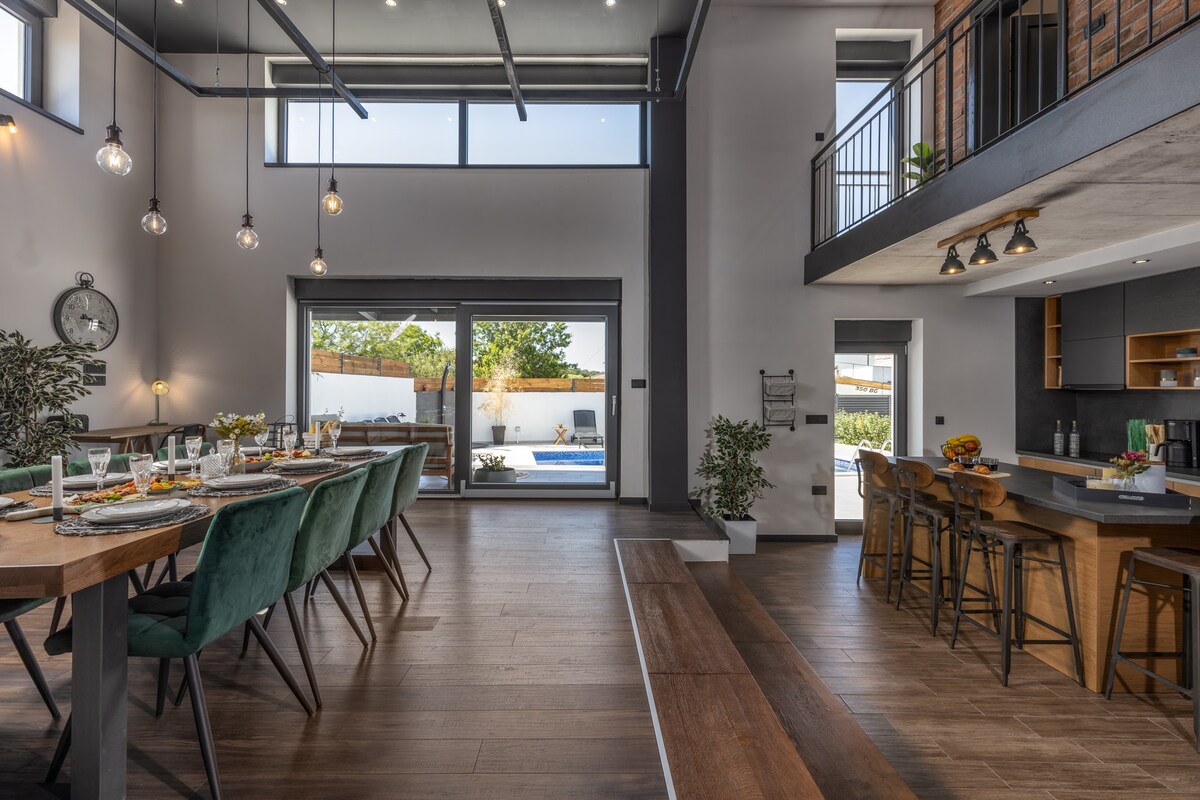 A spacious dining area features a long wooden table set with elegant dinnerware. Tall windows allow natural light to fill the space, with a view of the outdoor pool visible through sliding glass doors. Modern bar stools complement a sleek kitchen area in the background.