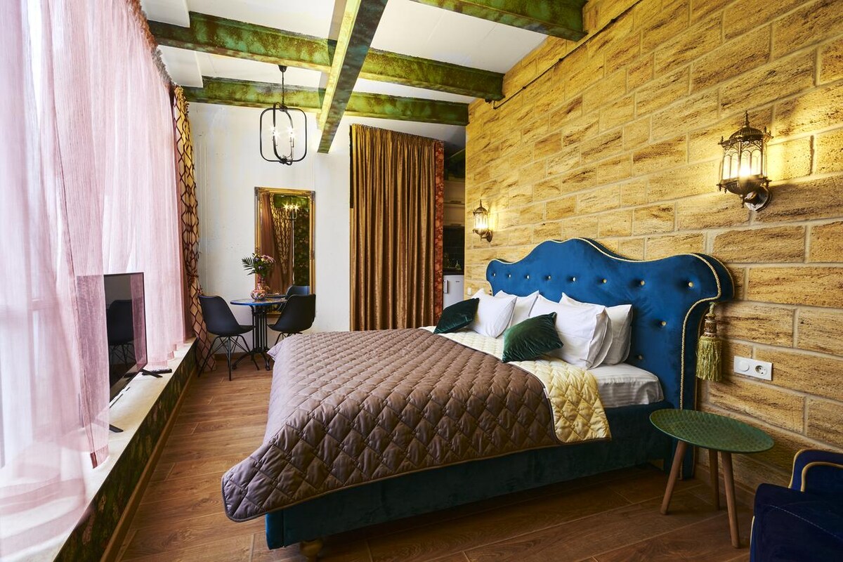 A spacious room is featured, with a large bed adorned with a textured quilt. The walls are textured with warm stone and accentuated by decorative lighting. Soft pink curtains frame a window, while a dining area with modern furniture is visible in the background.