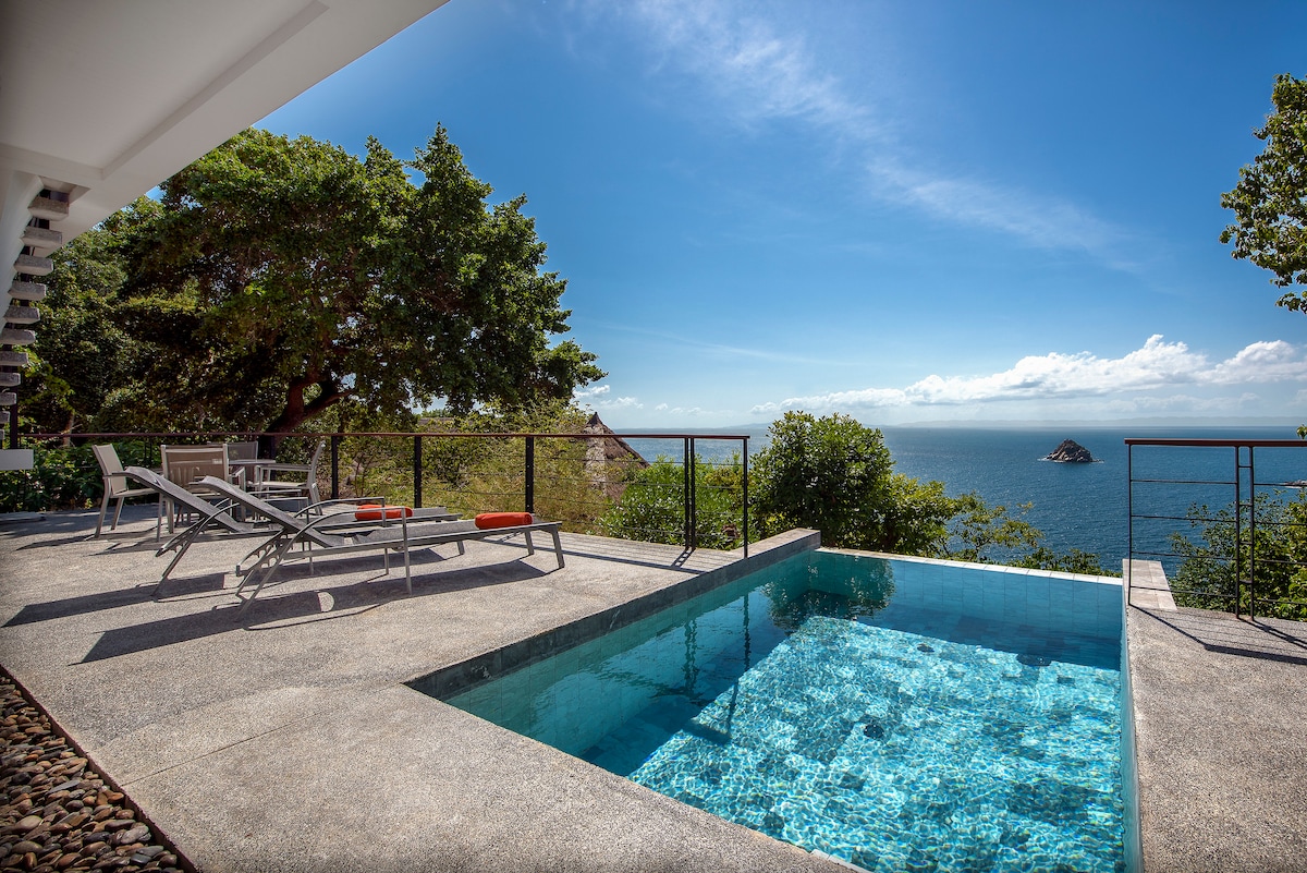 A private terrace features a small, clear pool surrounded by a smooth concrete deck. Comfortable lounge chairs are positioned for ocean views, with lush greenery framing the scene. The horizon displays a serene expanse of water under a bright blue sky.