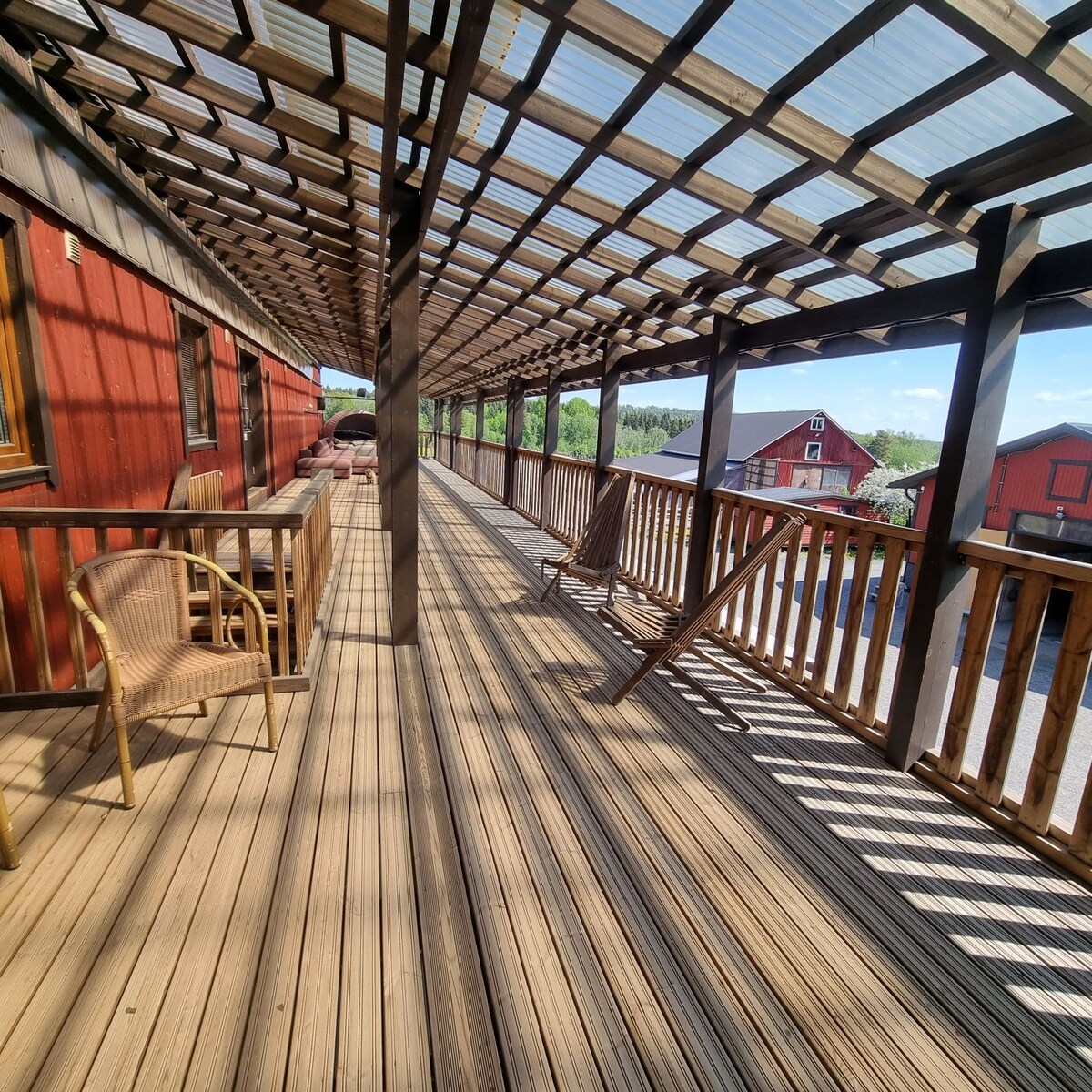 An expansive wooden terrace is covered with a transparent roof, allowing natural light to filter through. Several seating arrangements are available, including a wicker chair positioned against the railing. The view reveals a rural landscape with nearby red buildings visible in the distance.