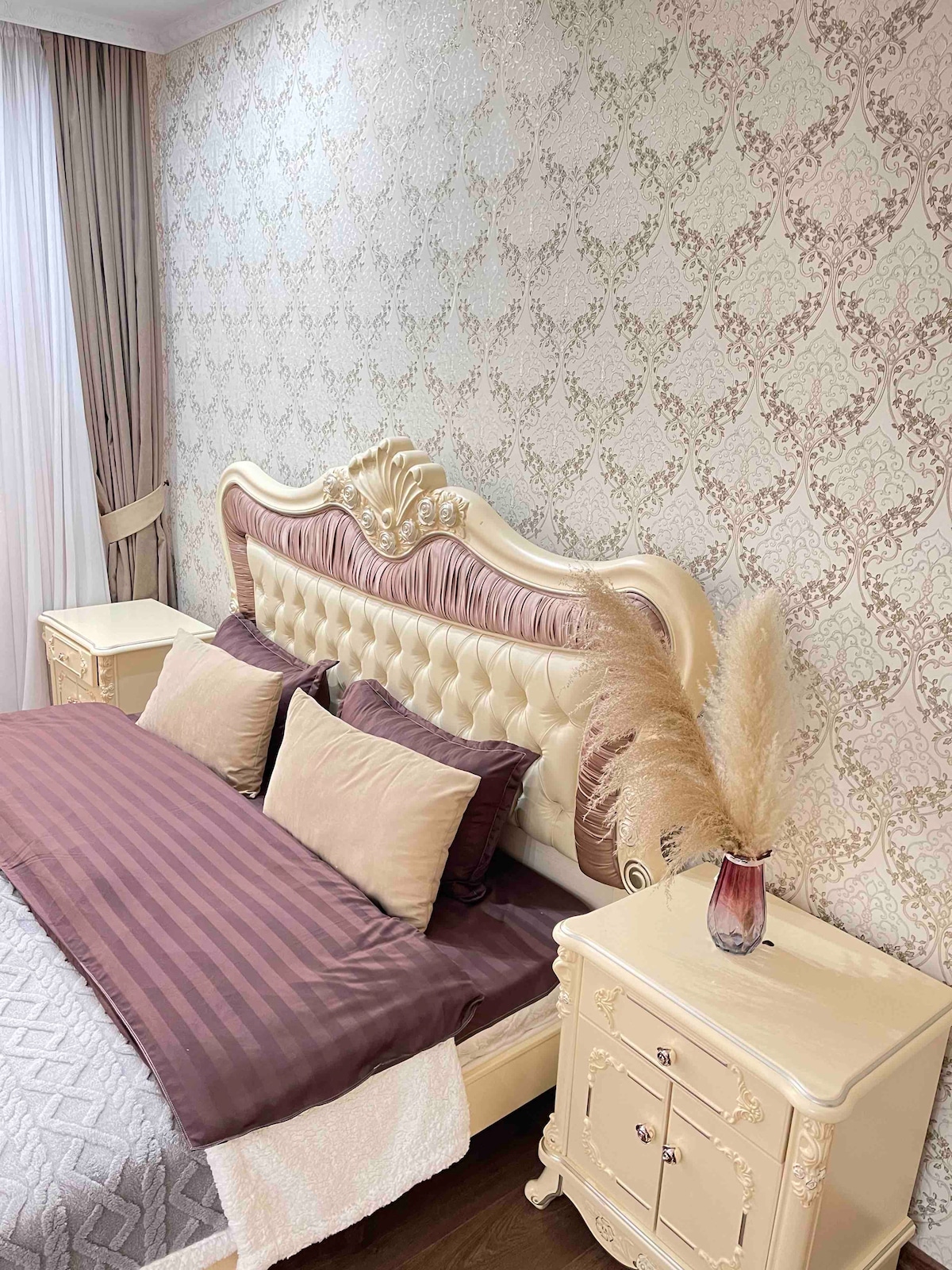 A cozy sleeping area features a tufted headboard in soft cream tones, complemented by decorative pillows in muted colors. The wall showcases an intricate pattern, and a vase with pampas grass sits atop one of the two nightstands.