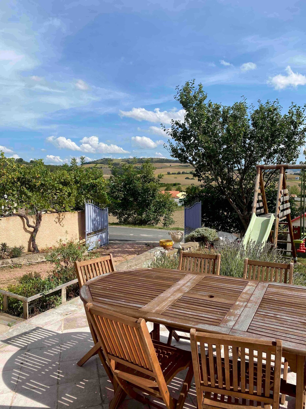 An outdoor terrace is featured with a wooden dining table surrounded by several matching chairs. In the background, lush greenery and rolling hills are visible under a clear blue sky, while a swing set adds a playful touch to the serene landscape.