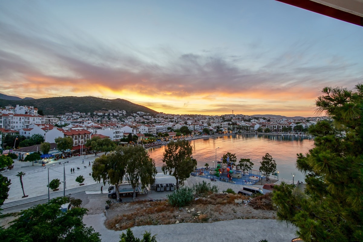 A scenic view captures the shoreline at sunset, with vibrant colors reflecting on the calm water. Waterfront buildings and greenery frame the image, while the distant hills provide a natural backdrop to the lively area filled with trees and public spaces.