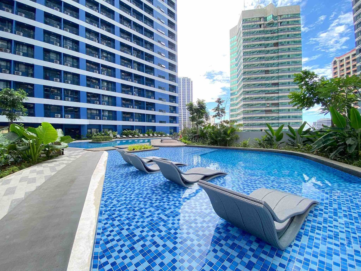 A serene pool area showcases blue mosaic tiles reflecting sunlight. Modern lounge chairs are positioned along the water's edge, surrounded by lush greenery and tropical plants. The backdrop features multi-story residential buildings, creating an inviting and refreshing outdoor space.