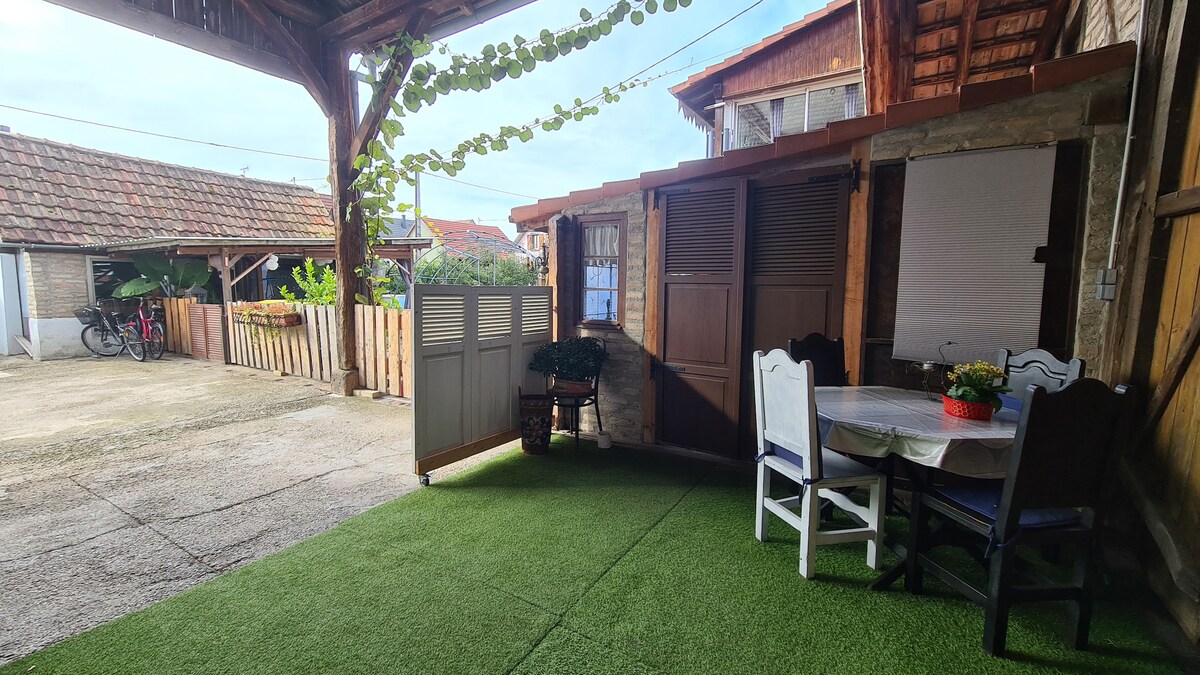 An outdoor seating area is highlighted, featuring a round table surrounded by four chairs. Lush green grass is laid underfoot, providing a comfortable space. In the background, rustic wooden structures and a small garden area contribute to the setting's charm.